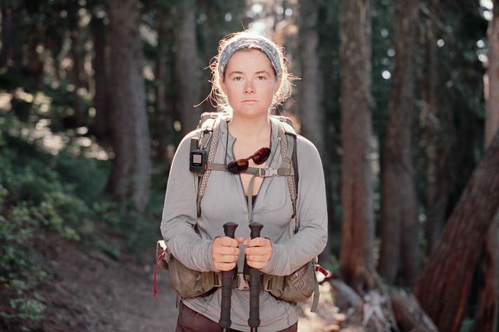 A woman wearing a backpack and holding trekking poles stands on a trail in a forest.