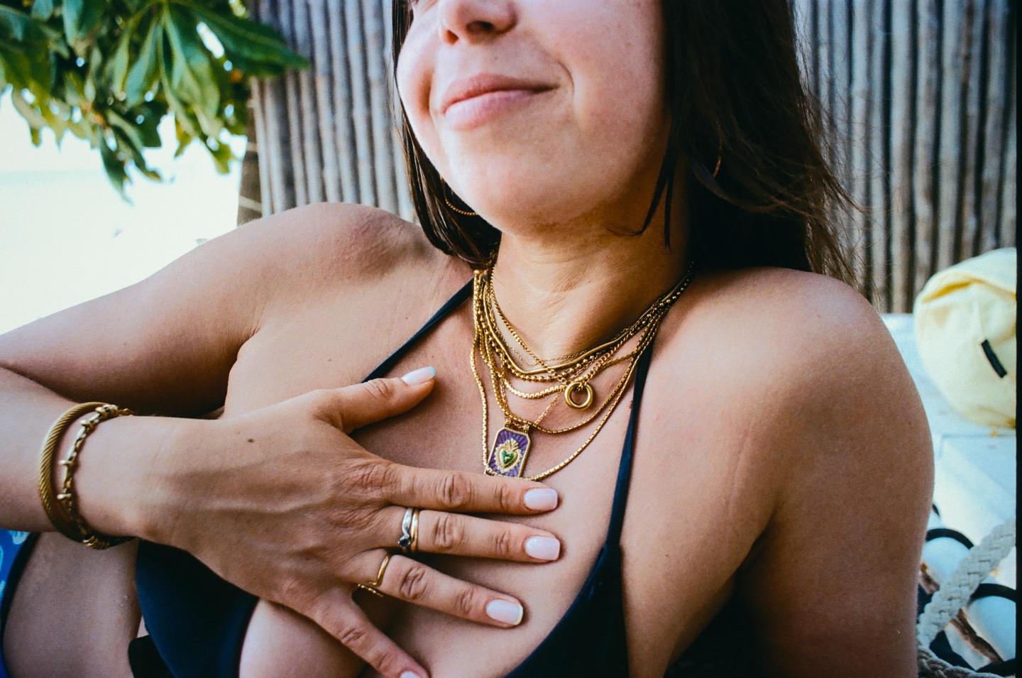 A woman wearing multiple gold necklaces and rings smiles while holding her hand to her chest.