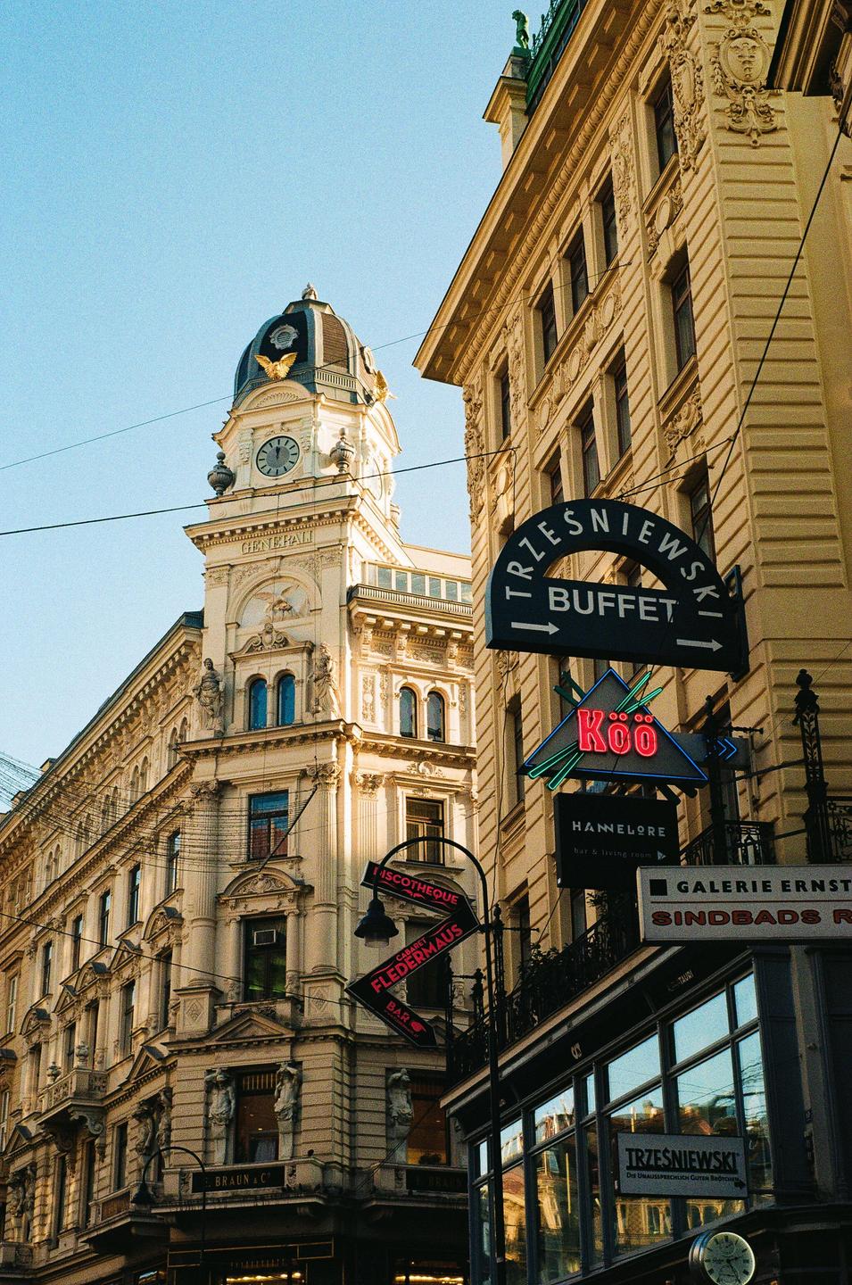 A tall, ornate building with a clock tower stands next to another building with several signs, including one for "Trzesniewski Buffet" and a neon sign for "Koo."