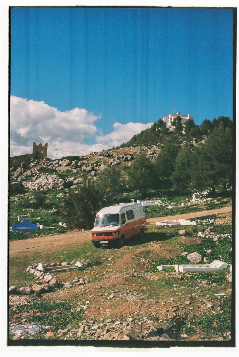 A white and orange van is parked on a dirt road in a hilly landscape with a building on a distant hilltop.