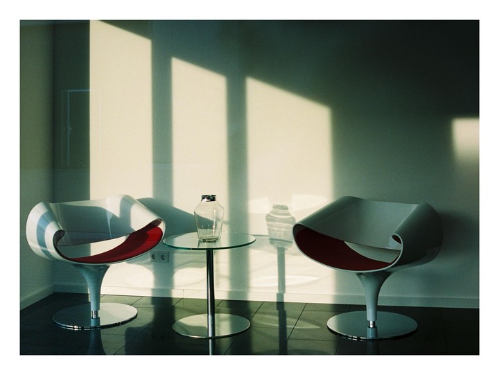 Two modern chairs with red seats flank a glass table with a vase, all illuminated by strong window light and shadows.