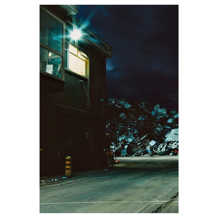 A building with a brightly lit window stands next to a large pile of scrap metal under a dark night sky.