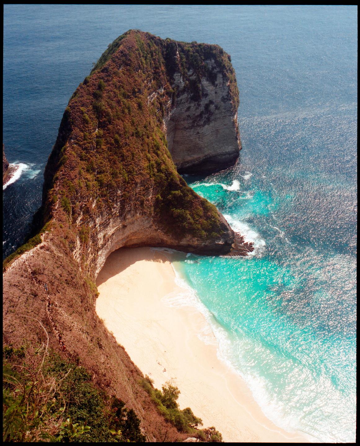 A high-angle view shows a white sand beach and turquoise water at the base of a large, green-covered cliff.