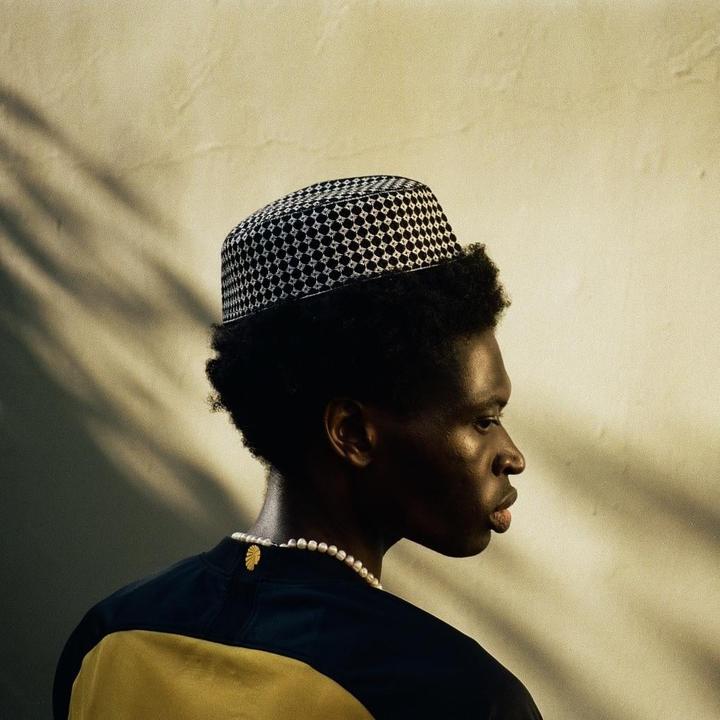 A man wearing a patterned hat and a pearl necklace is shown in profile against a light wall with shadows.