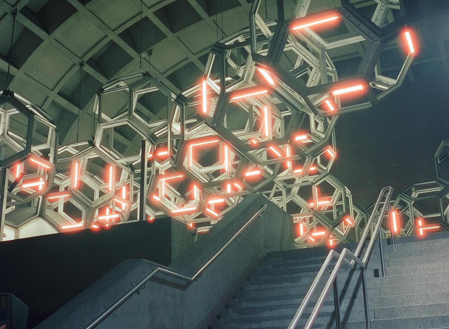 A concrete staircase with metal railings leads up towards a ceiling adorned with geometric light fixtures.