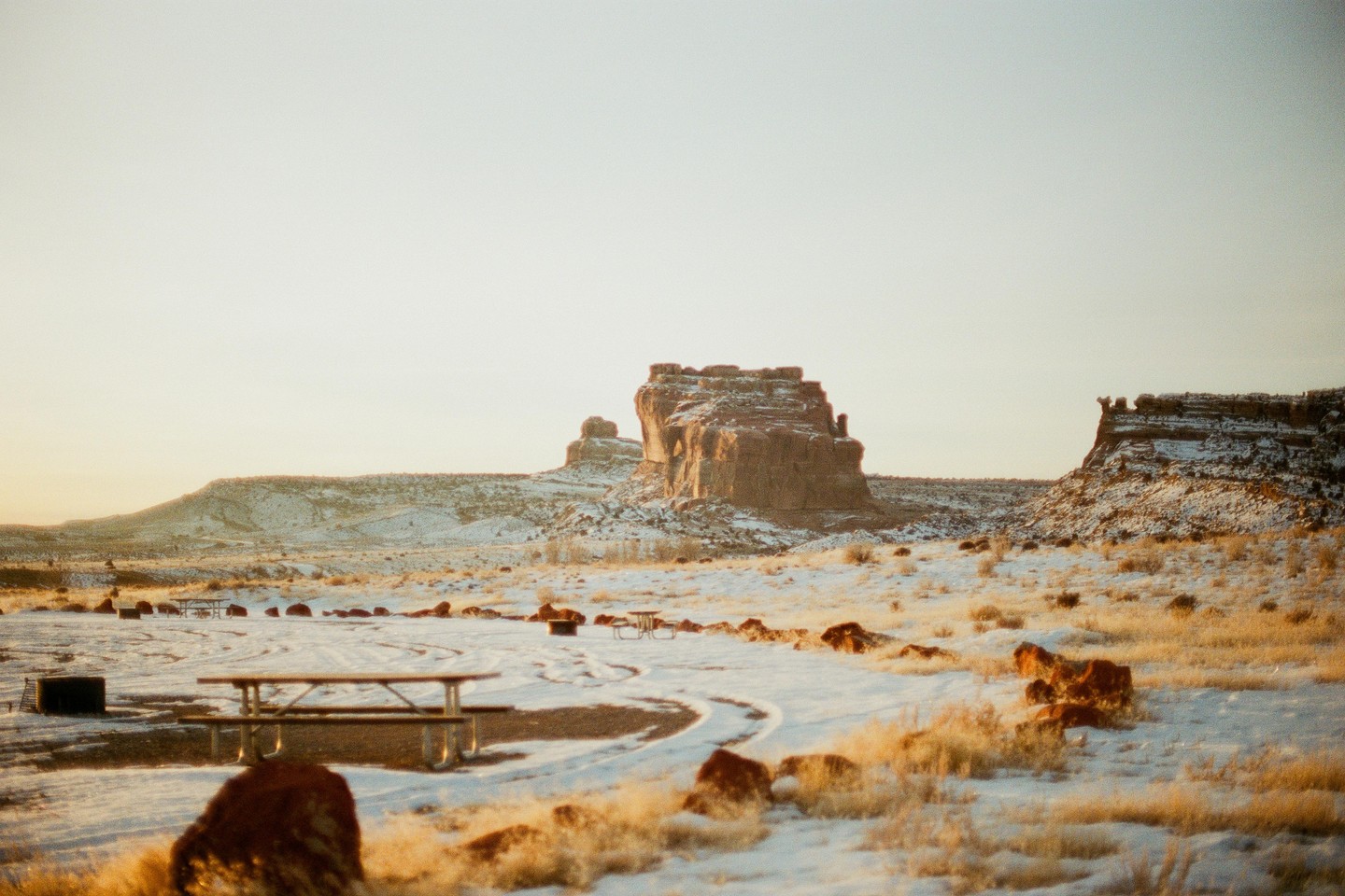 A snowy desert landscape features mesas and picnic tables under a bright sky.