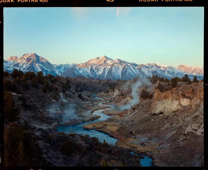 A winding river flows through a valley with steam rising from hot springs, leading to snow-capped mountains under a clear sky.