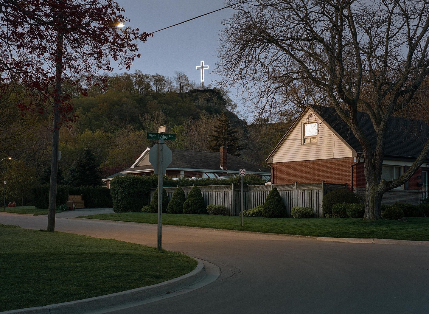 A glowing cross stands on a hill overlooking a residential street with houses and trees.