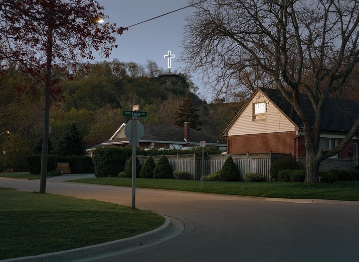 A glowing cross stands on a hill overlooking a residential street with houses and trees.