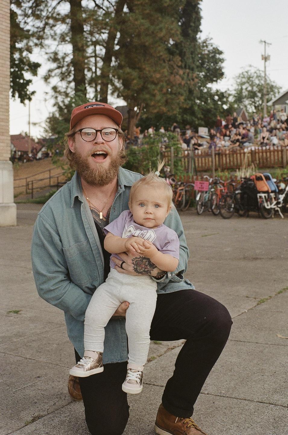 A man kneels while holding a baby with a crowd of people in the background.