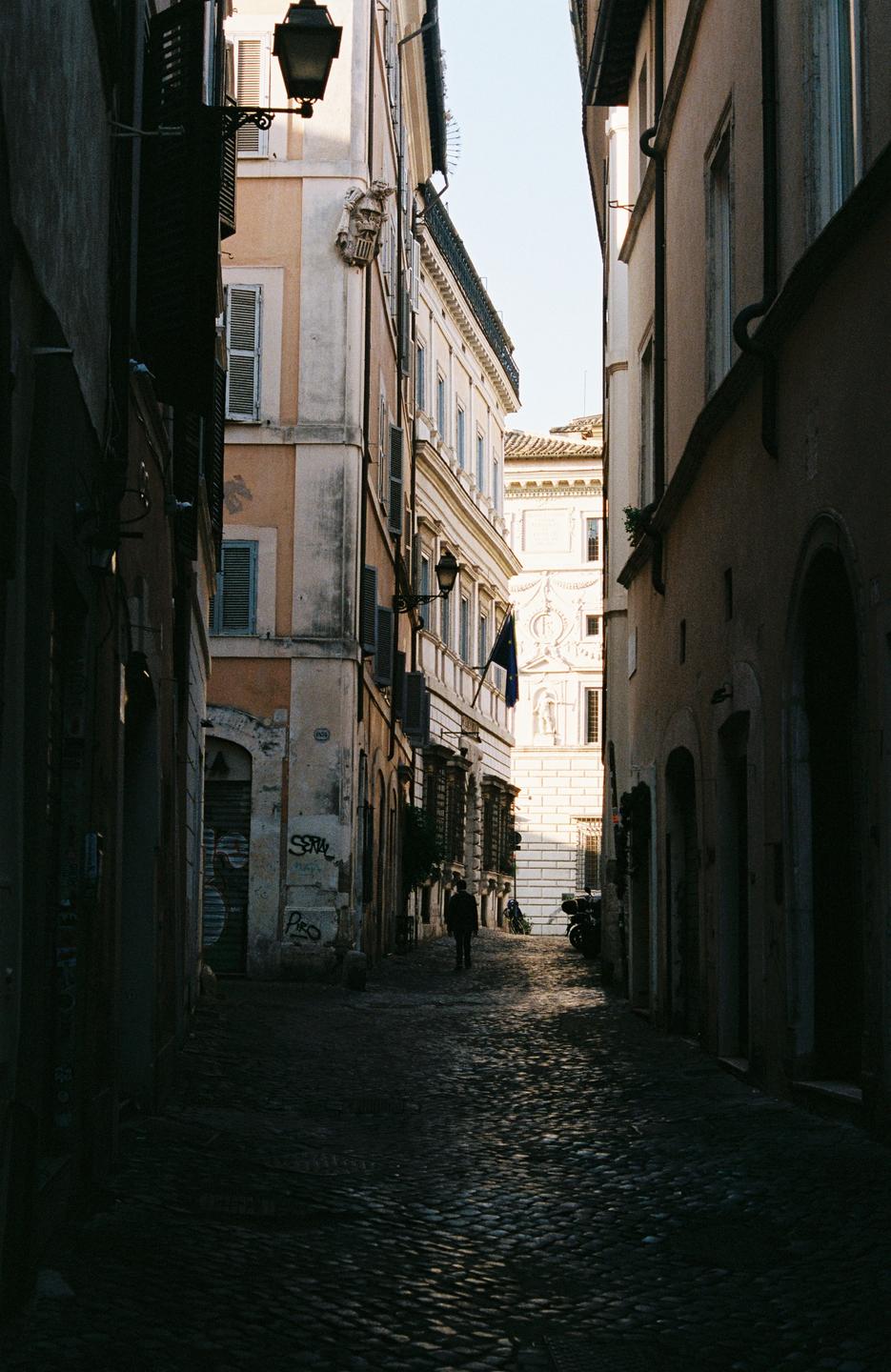 A person walks down a narrow, shadowed cobblestone alley lined with tall buildings.