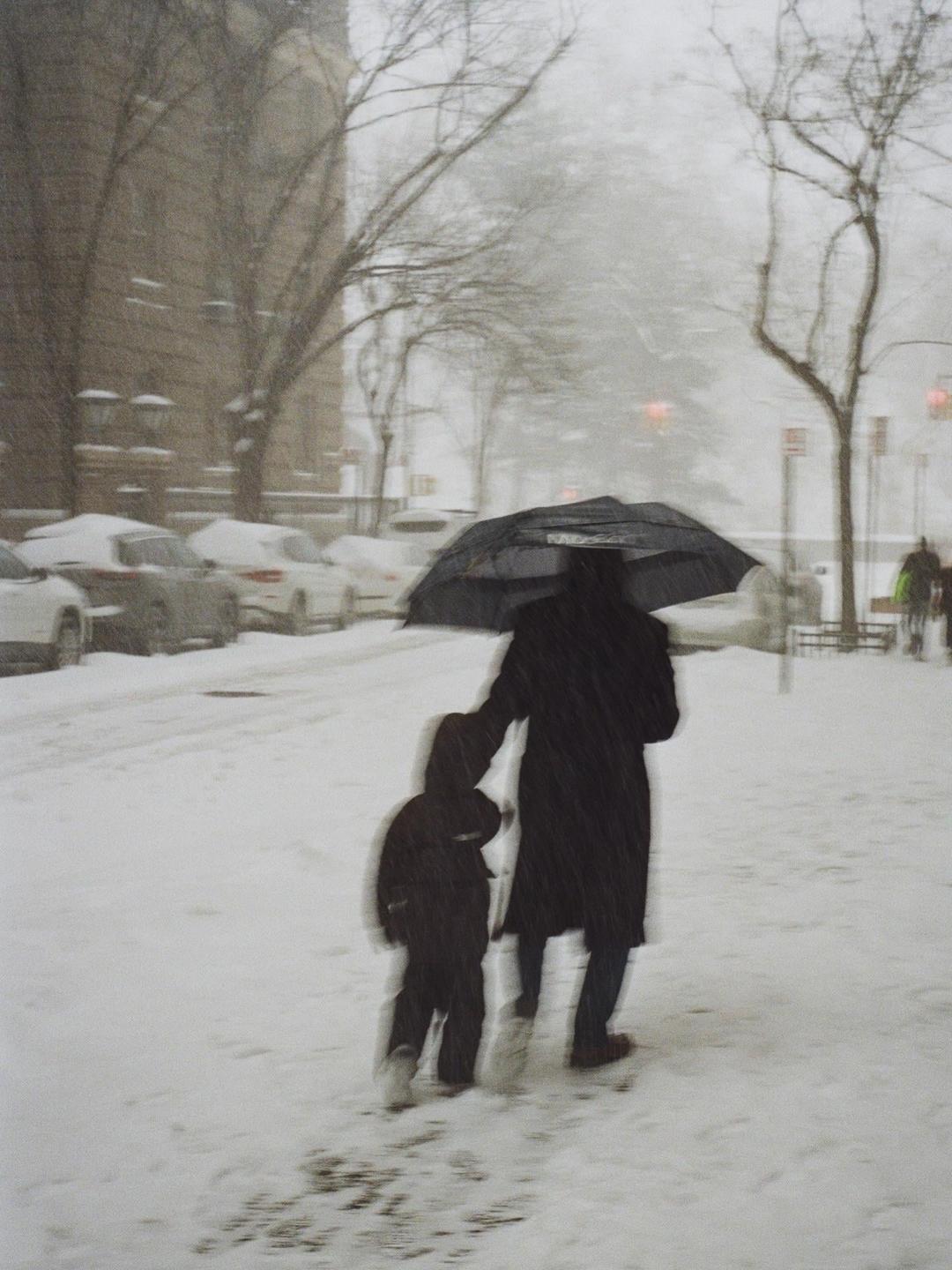 A person holding an umbrella walks with a child through a snowy street.