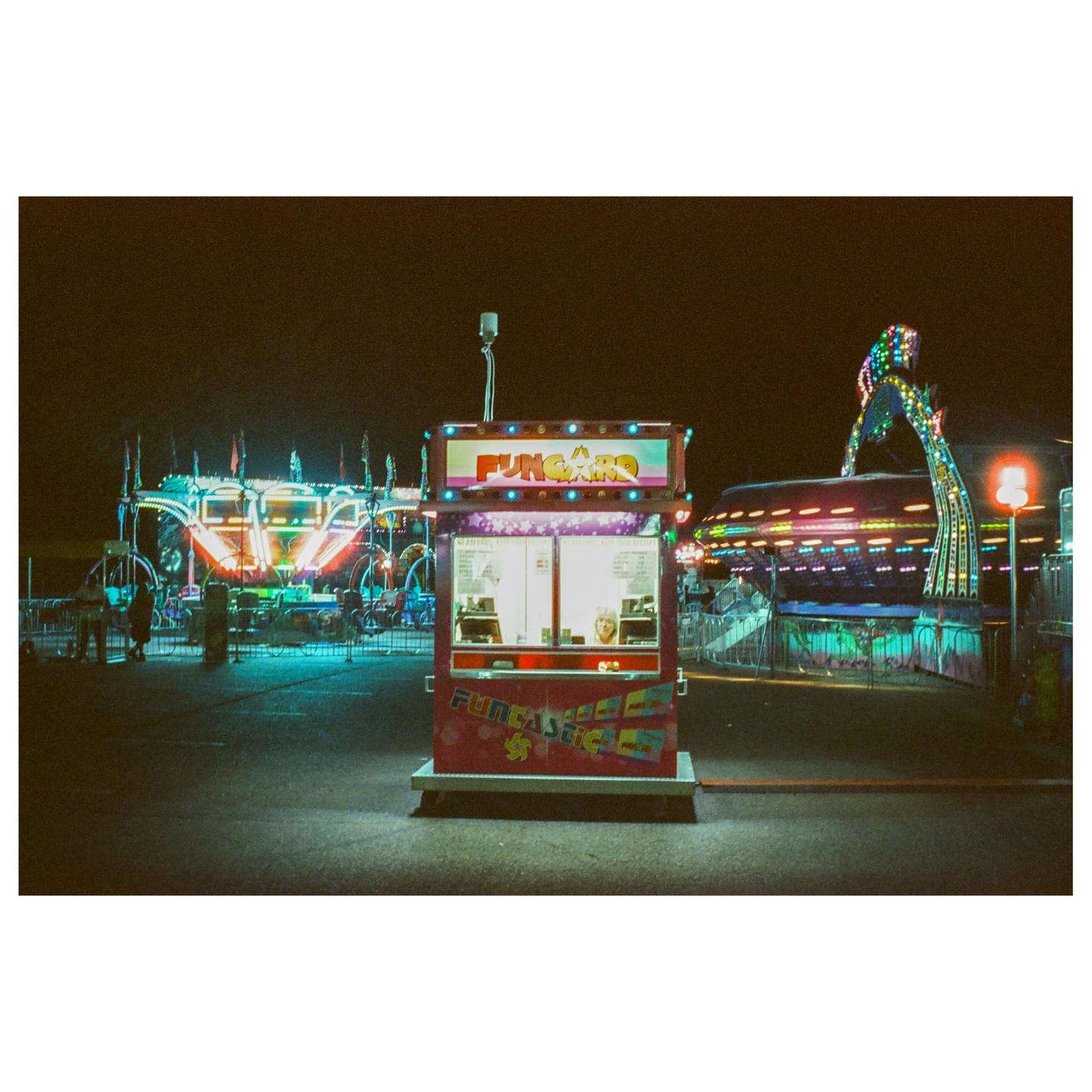 A brightly lit ticket booth stands in the foreground of a carnival at night, with various rides visible in the background.