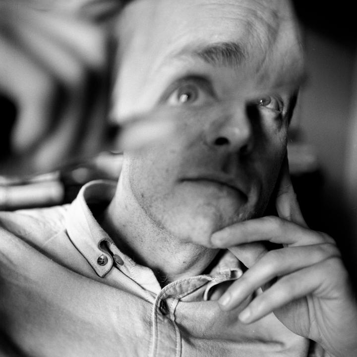 A man with a hand on his chin looks up and to the right, with a distorted reflection of his face visible in the upper left.
