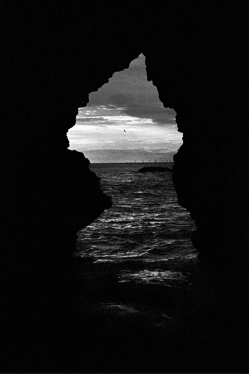 A dark rock formation frames a view of the ocean with a cloudy sky and a distant pier.