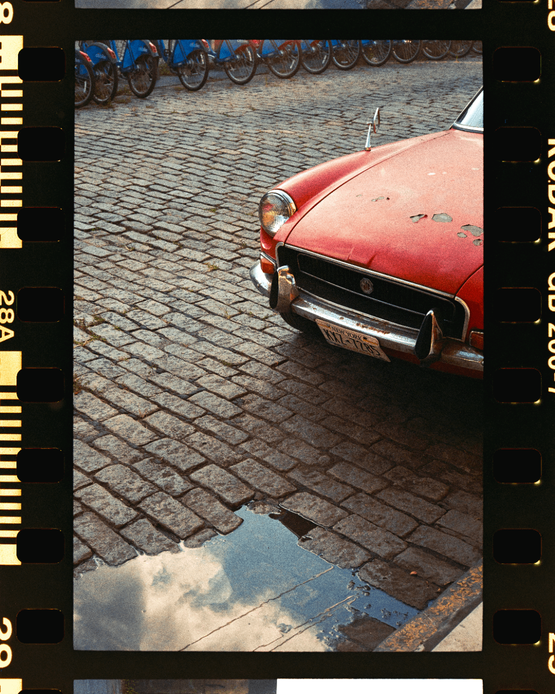 A red car is parked on a cobblestone street with a puddle reflecting the sky.
