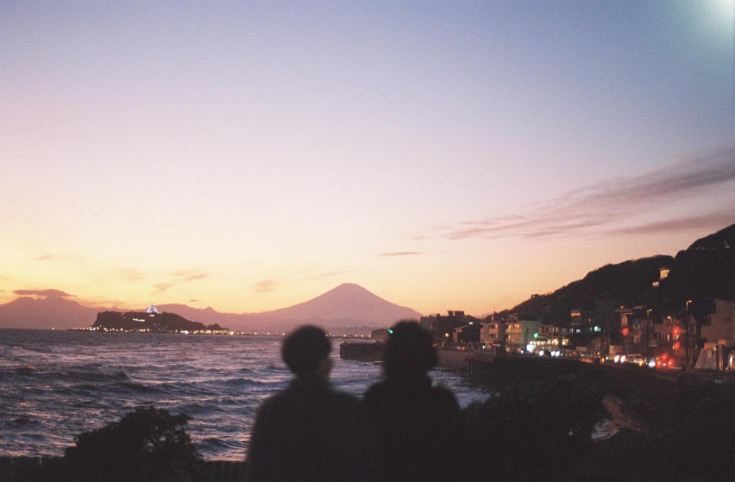 Two people look out at a body of water with a mountain and a town in the distance at dusk.