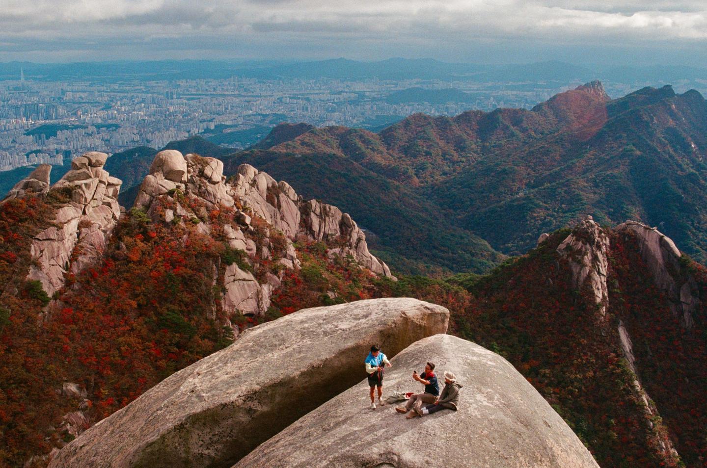 Three people are on a large rock formation overlooking a city and mountains with autumn foliage.
