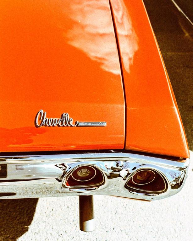 The rear of an orange Chevelle car is visible, showing the chrome bumper and exhaust pipe.