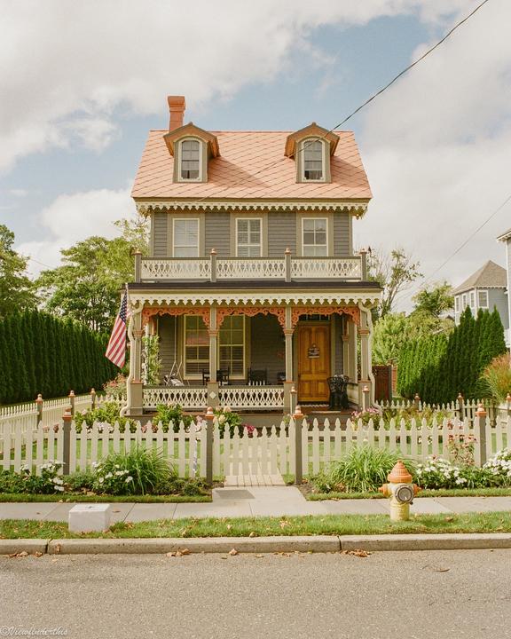 A charming Victorian house with a pink roof and a white picket fence stands under a partly cloudy sky.