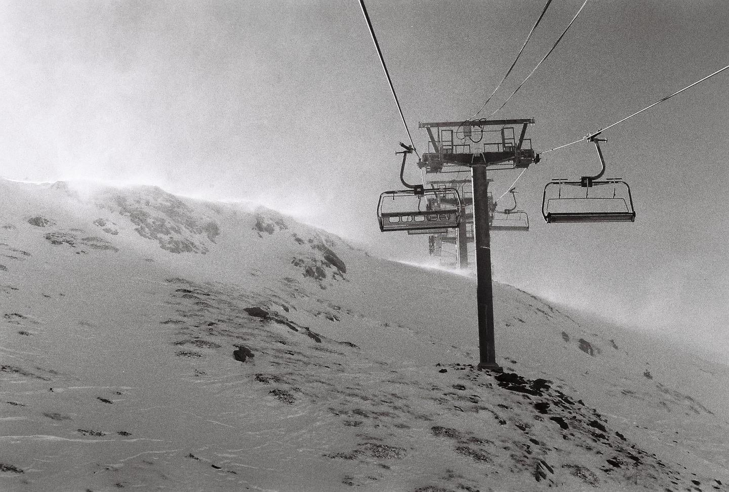 A ski lift ascends a snow-covered mountain with blowing snow.