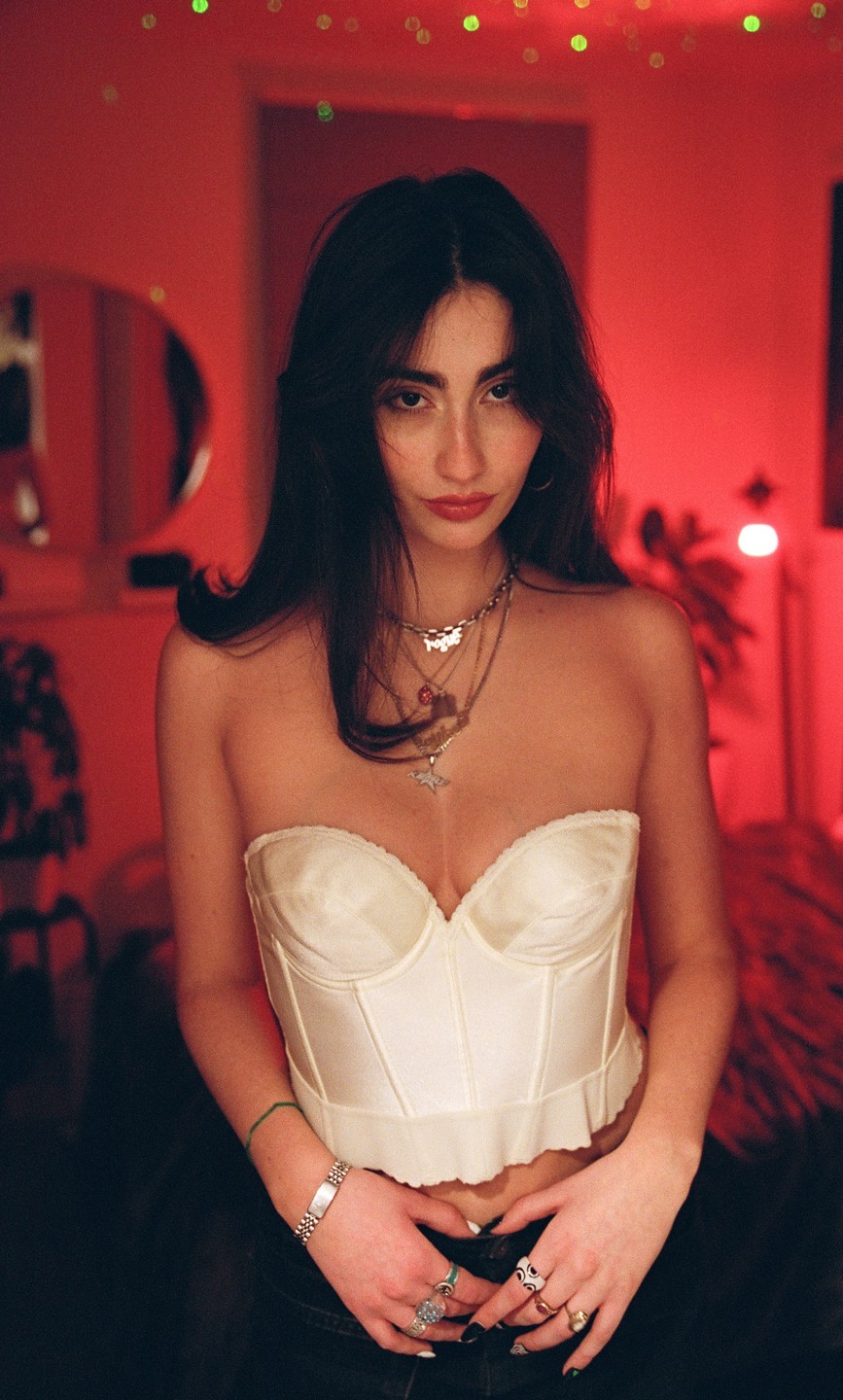A woman wearing a white corset and multiple necklaces stands in a room lit with red light.