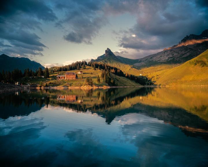 A large building with many windows sits on a grassy hill overlooking a lake, with mountains and clouds in the background.