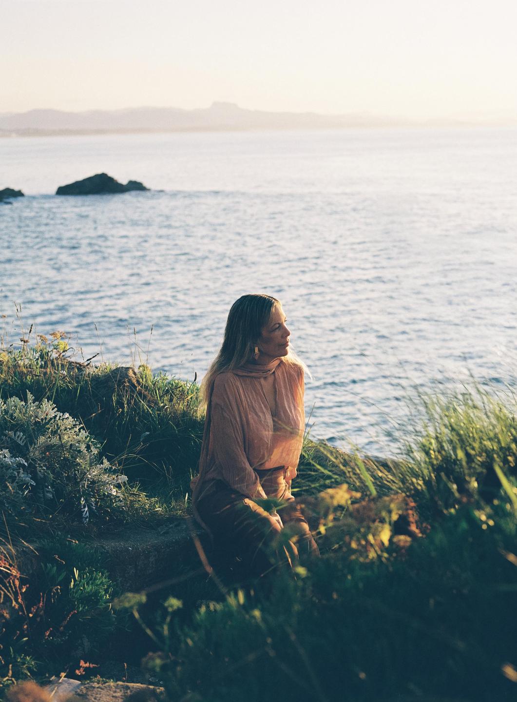 A woman sits on a grassy cliff overlooking the ocean at sunset.