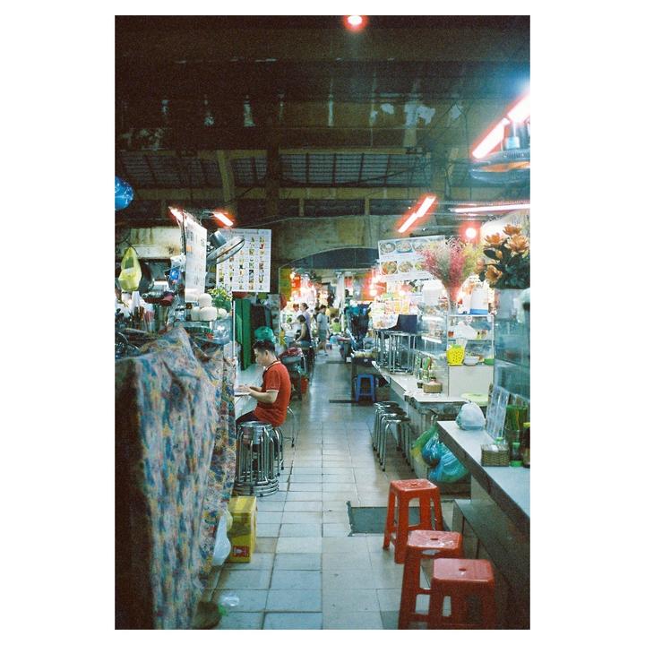 A man sits at a counter in a market with red stools in the foreground and other people in the background.