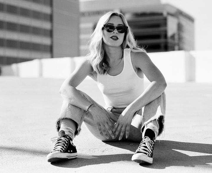 A woman wearing sunglasses, a tank top, and jeans sits on the ground with buildings in the background.