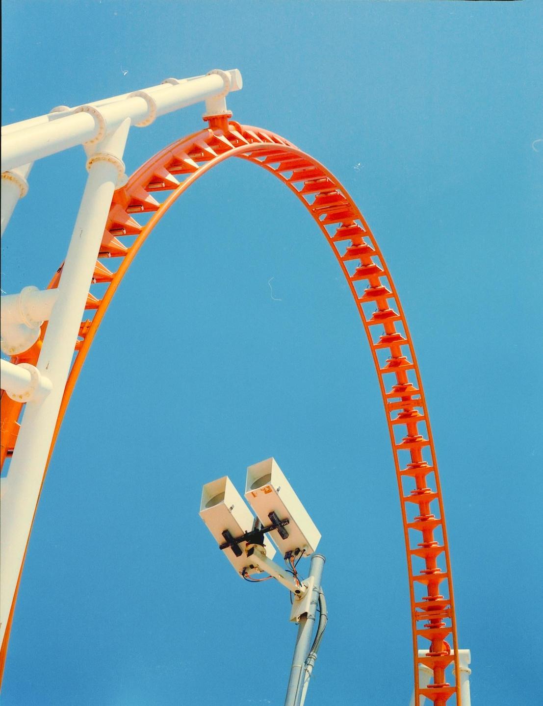 An orange and white roller coaster track curves against a clear blue sky, with two white security cameras mounted on a pole in the foreground.