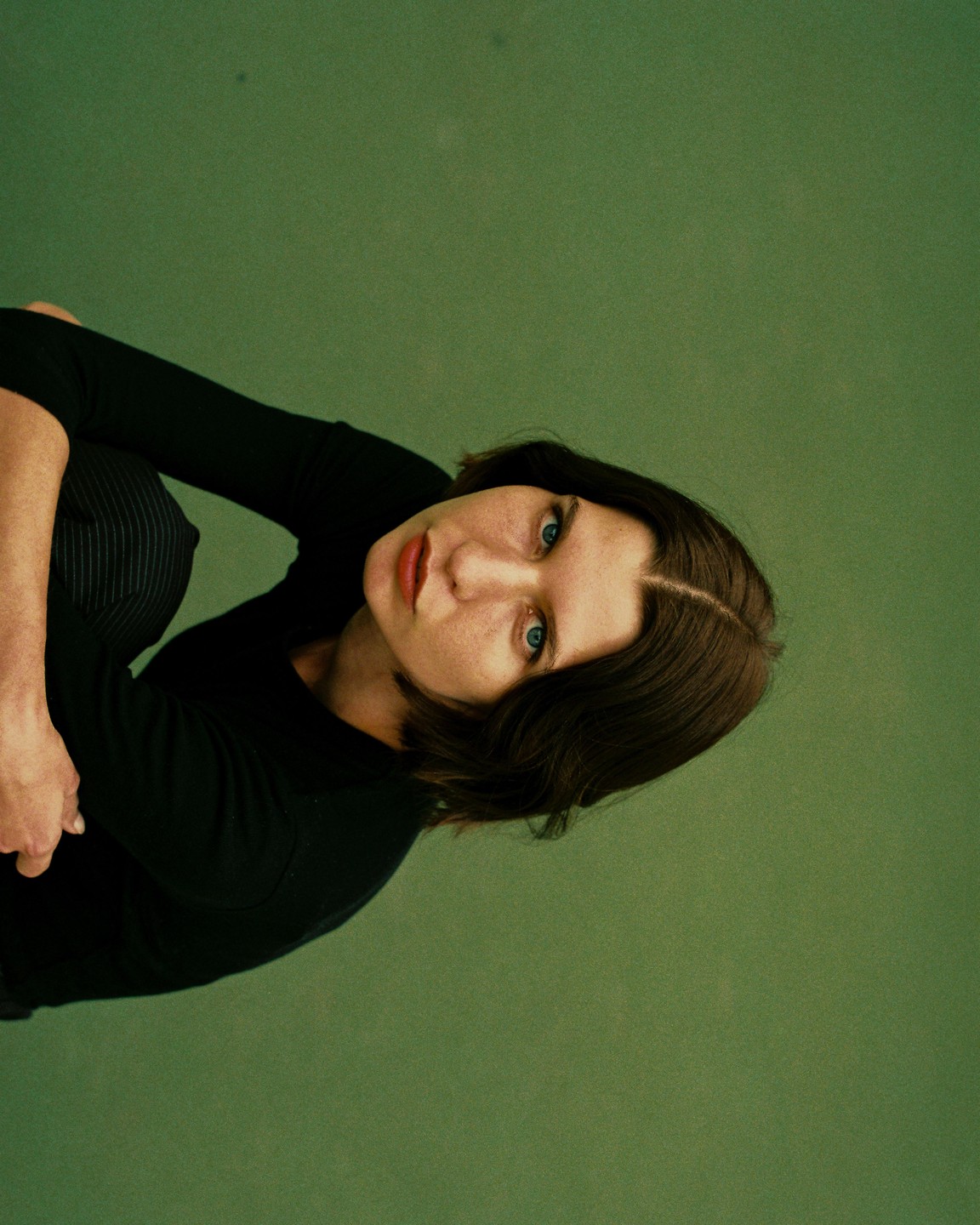 A woman with short brown hair and blue eyes looks up at the viewer against a green background.