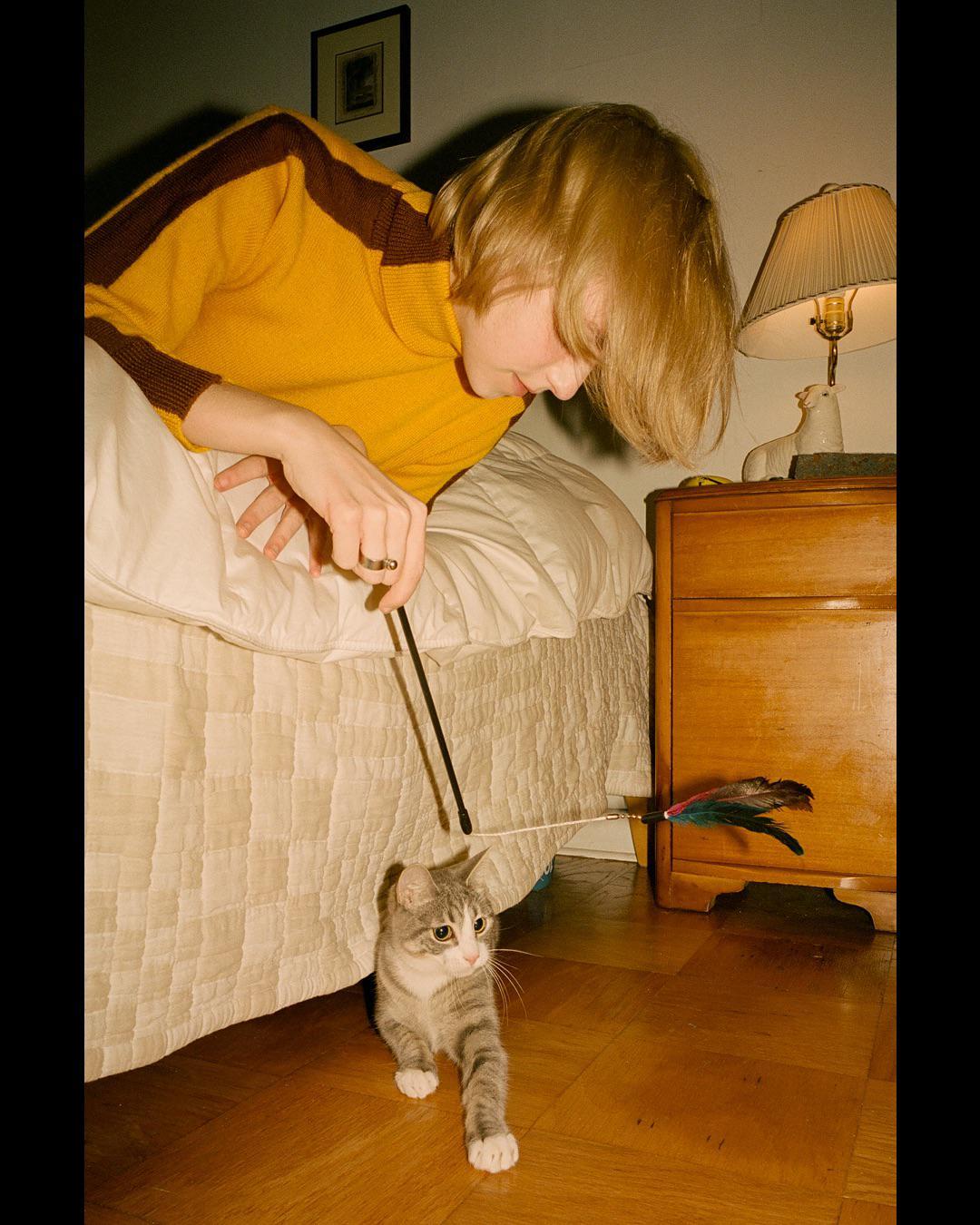 A woman plays with a cat using a feather toy next to a bed and a nightstand.