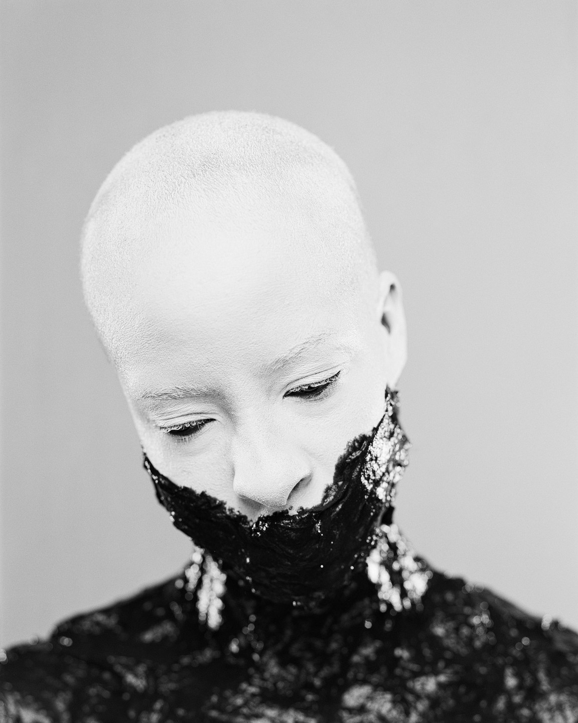 A person with a shaved head and white face paint looks down, with black paint covering their mouth and neck.