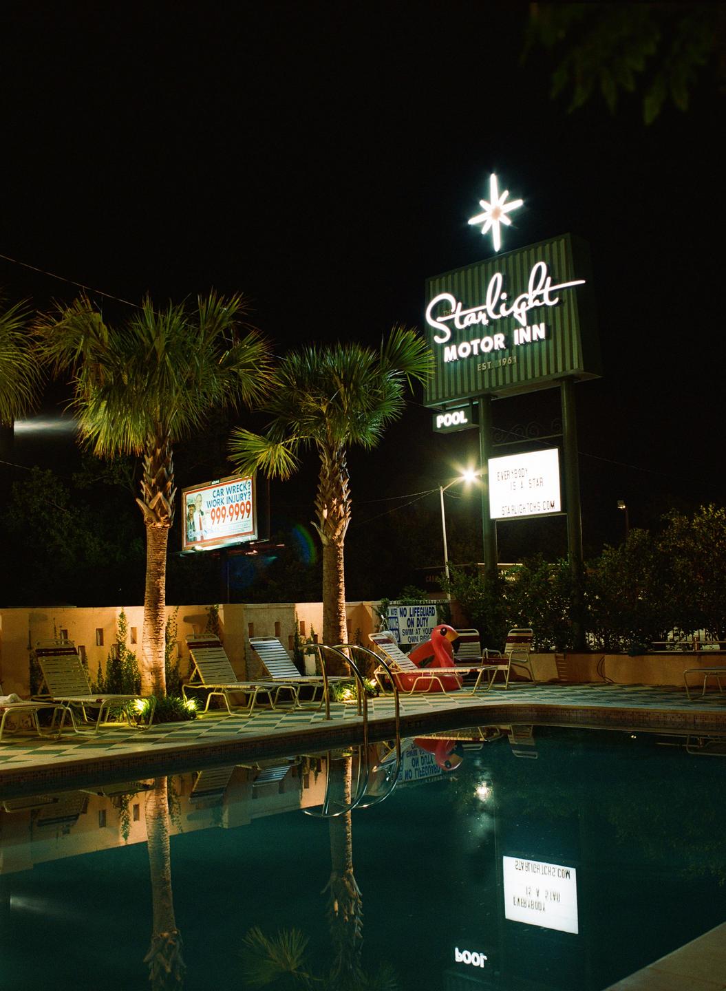 A neon sign for the Starlight Motor Inn stands above a swimming pool and palm trees at night.
