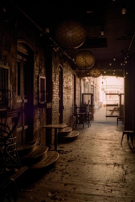 A narrow alleyway with brick walls, tables, and chairs is illuminated by string lights and large spherical lanterns.