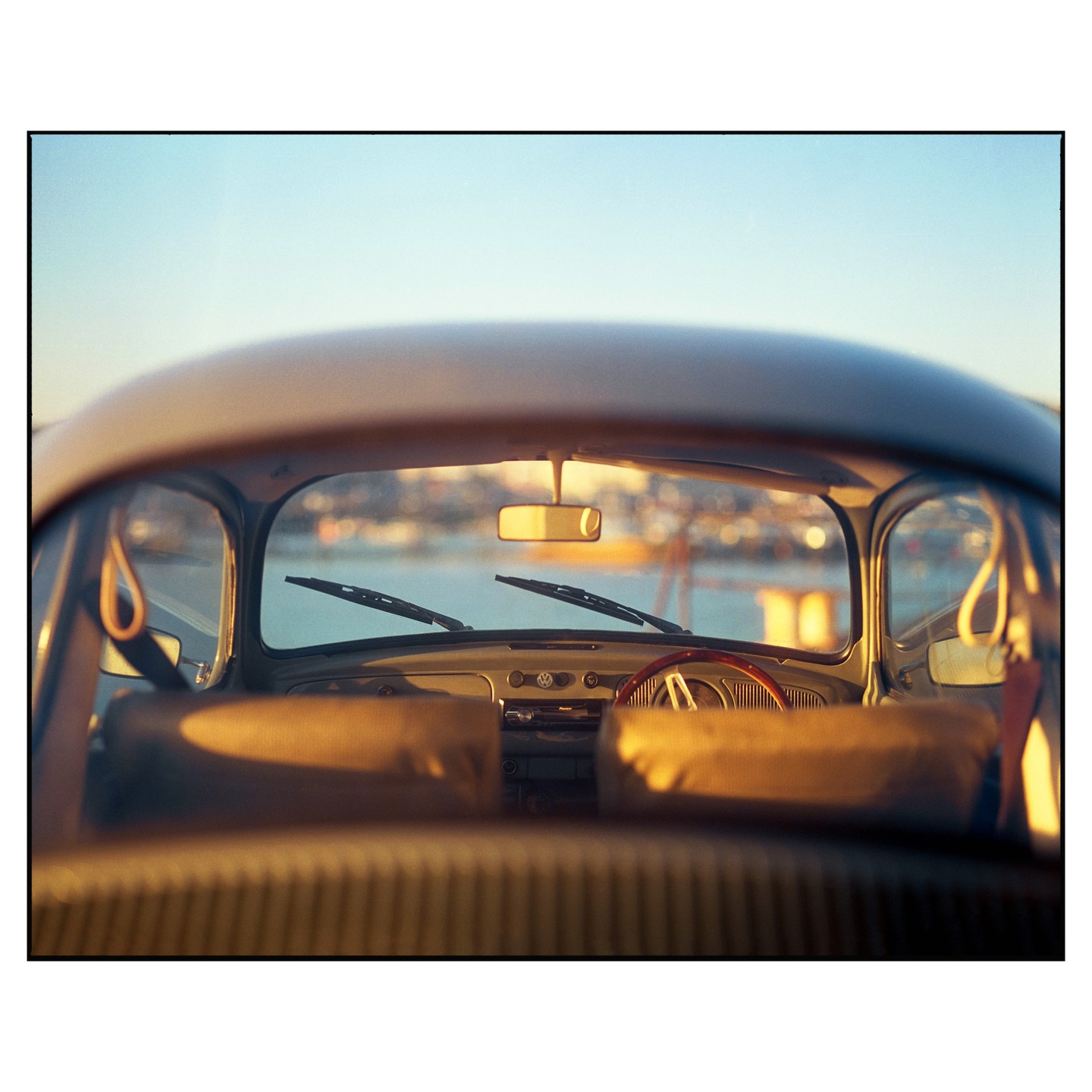 The interior of a vintage Volkswagen Beetle is visible through the rear window, with a body of water and a city in the background.