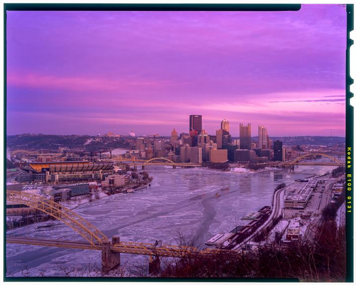 A cityscape with a frozen river and bridges is visible under a vibrant purple and pink sky.