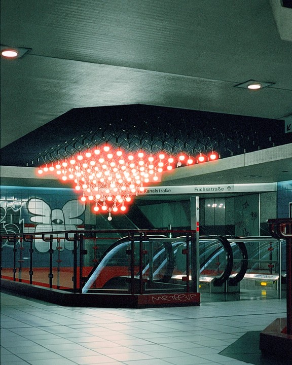 A red light fixture hangs above escalators in a subway station with graffiti on the walls.