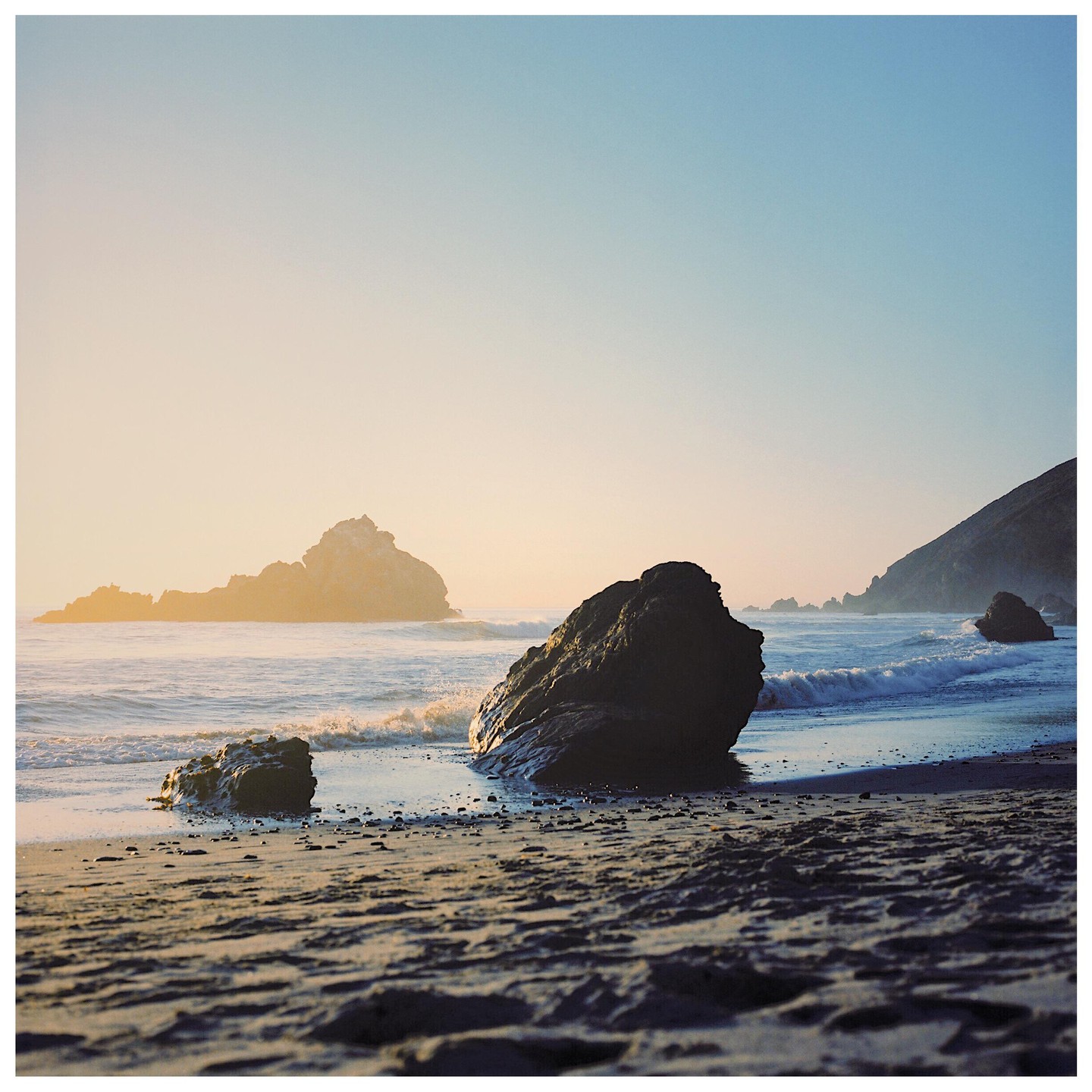 Waves crash on a sandy beach with large rocks and distant hills under a clear sky.