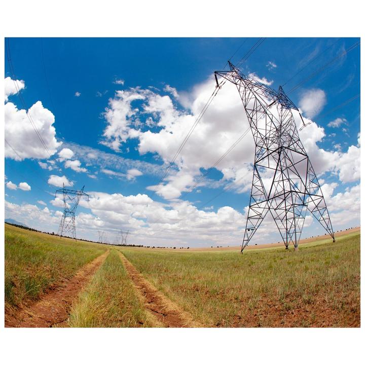 A dirt path leads through a grassy field with power lines and pylons under a blue sky with white clouds.