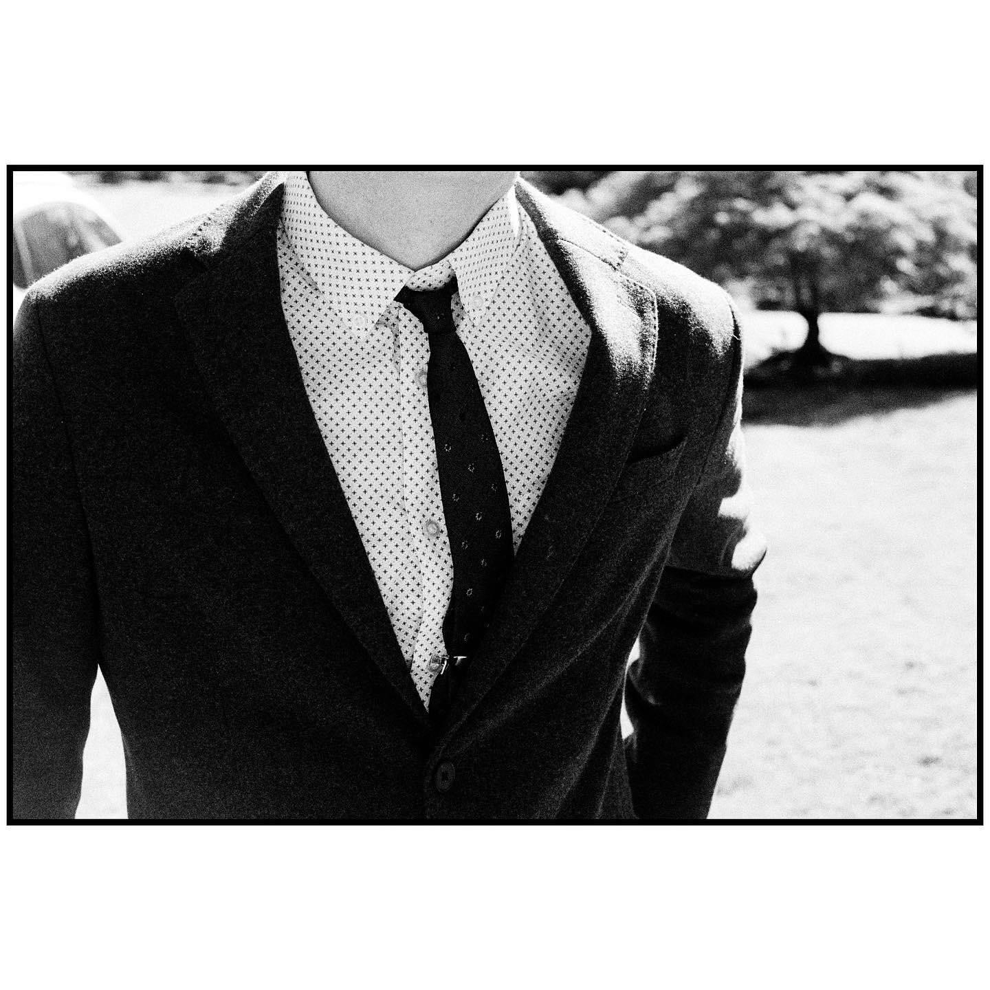 A man wears a suit jacket, a patterned shirt, and a tie with a tie clip, standing in a grassy outdoor area with a tree in the background.
