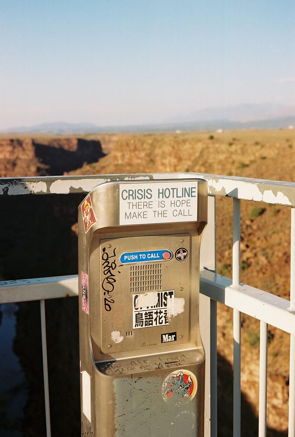 A crisis hotline call box with graffiti and stickers is mounted on a railing overlooking a canyon.
