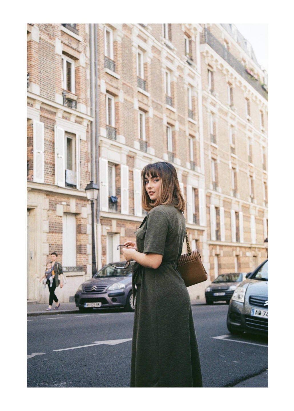A woman in a green dress stands on a city street looking over her shoulder, with buildings and cars in the background.