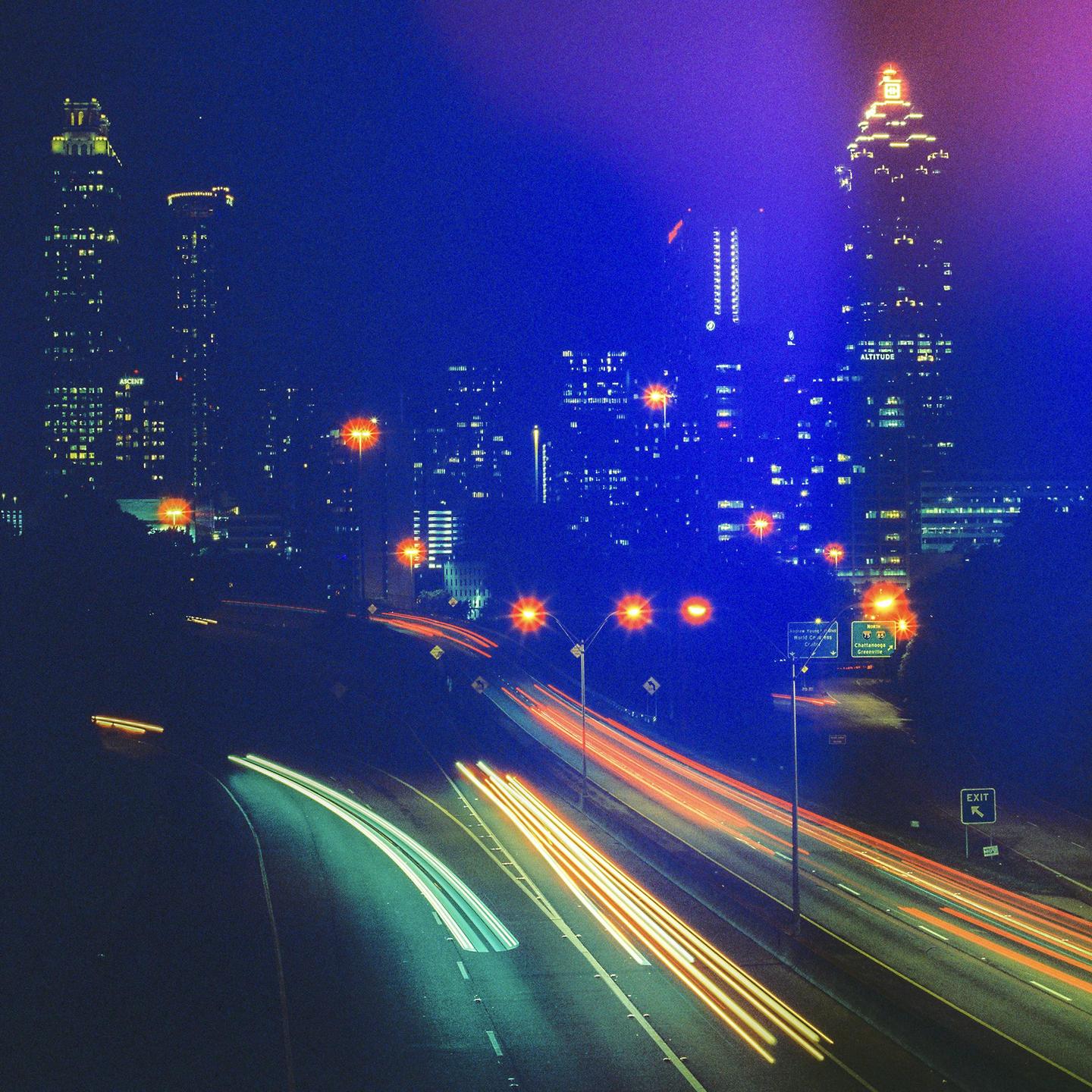 A long exposure captures light trails from vehicles on a highway at night with a city skyline in the background.