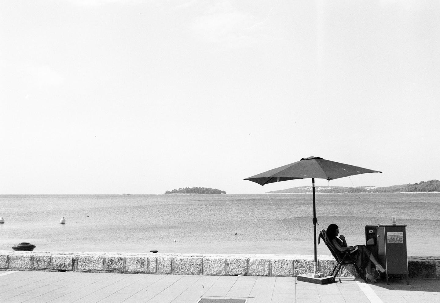 A woman sits under a large umbrella next to a body of water with an island in the distance.