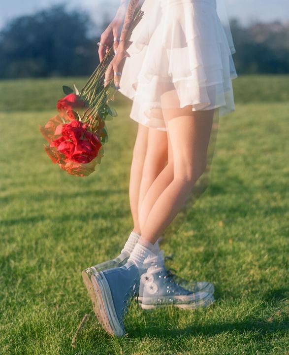 A woman in a white dress and blue sneakers holds a bouquet of red roses in a grassy field.