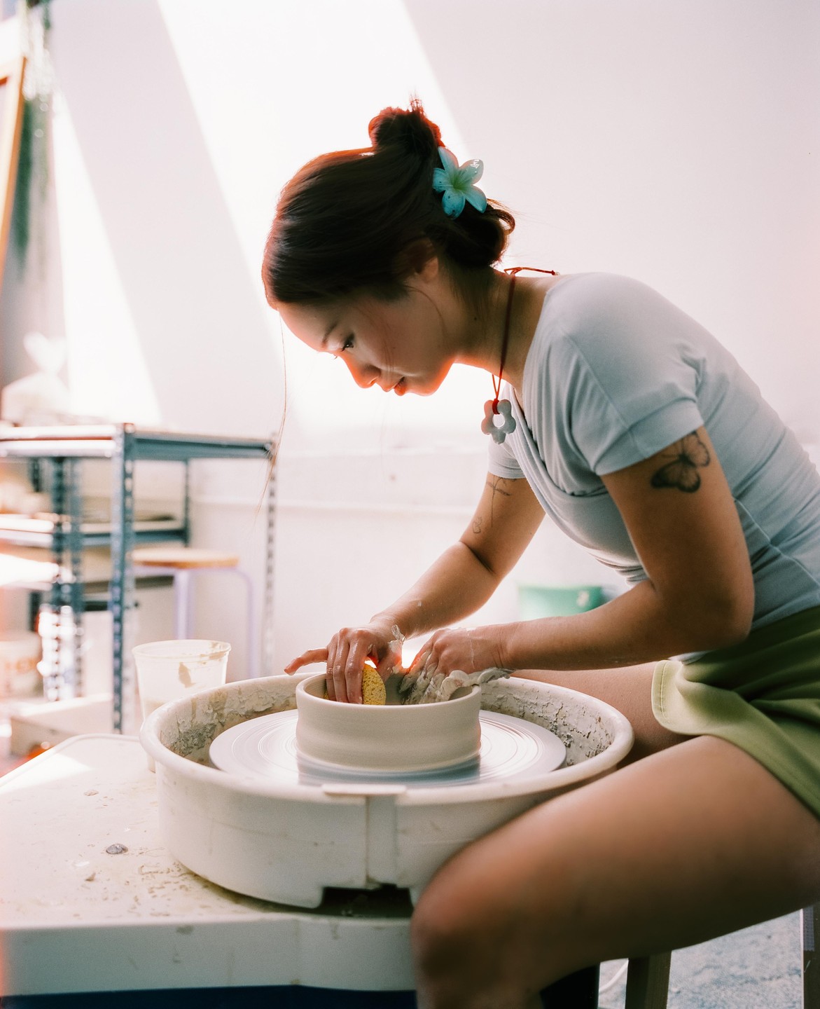 A woman with a flower in her hair is working on a pottery wheel in a bright studio.