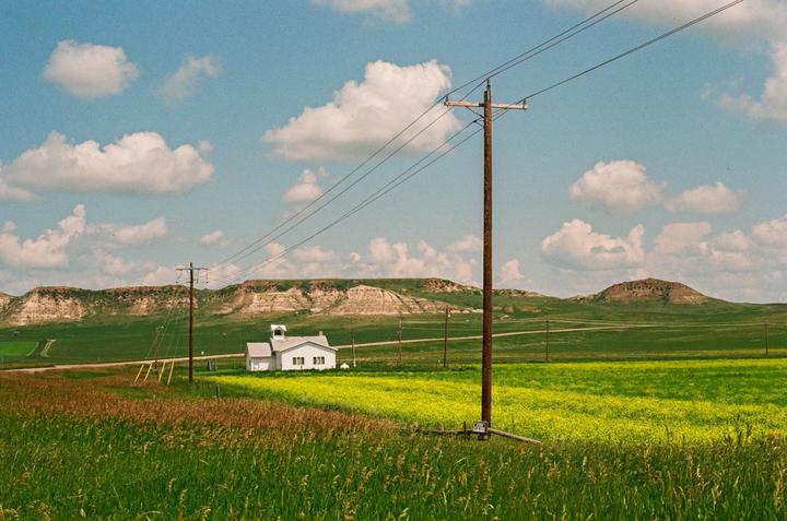 A white schoolhouse stands in a green field with yellow flowers, under a blue sky with clouds, with hills and power lines in the background.