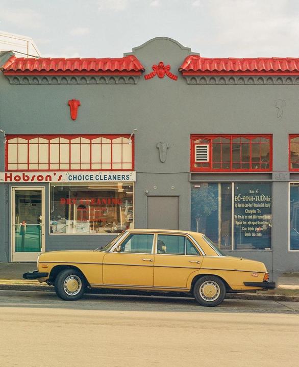 A yellow car is parked on a street in front of a gray building with red trim.
